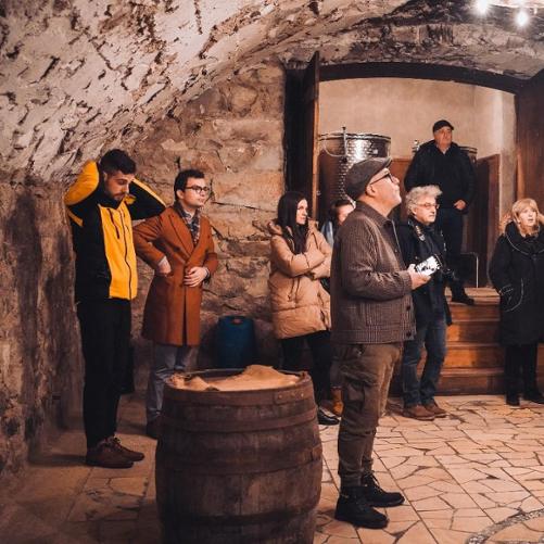 Dziesięcioro, ciepło ubranych osób zwiedza piwnicę z winami.