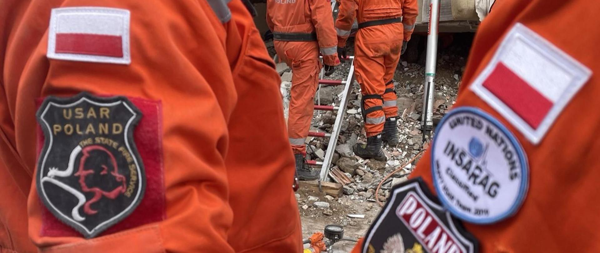 Na pierwszym planie emblematy na mundurach grupy poszukiwawczo-ratowniczej, w tle trzech ratowników w trakcie prowadzonych działań na gruzowisku