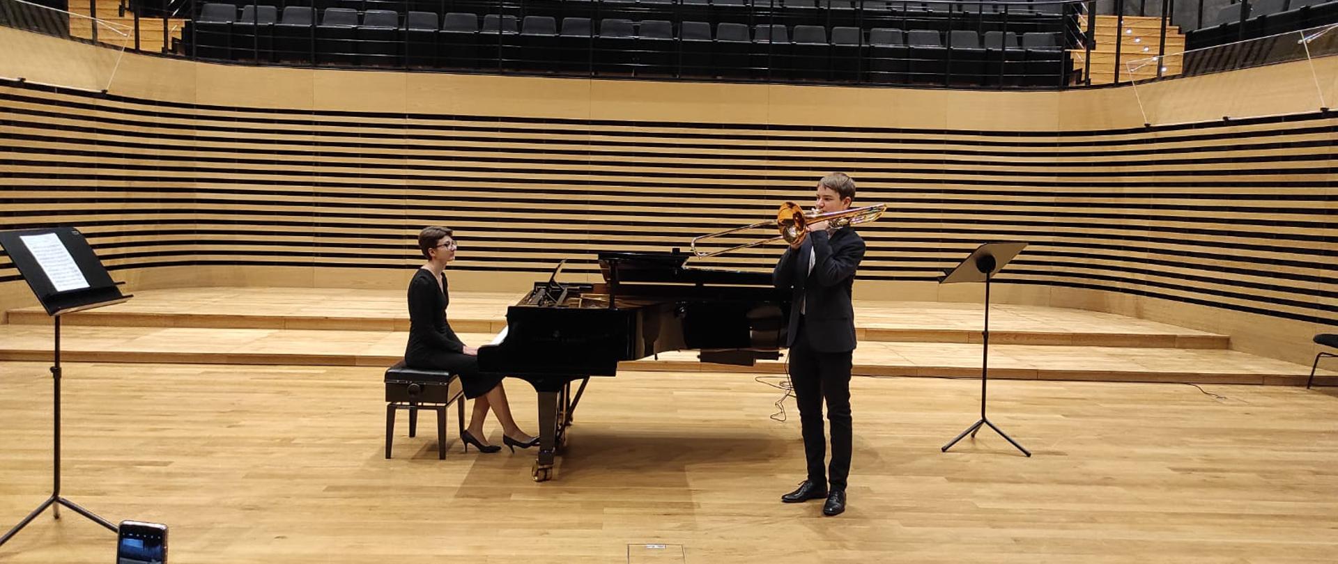 Zdjęcie przedstawia ucznia Damiana Kołodziejczaka na scenie w stroju galowym, trzyma puzon, przy fortepianie akompaniuje Agnieszka Unolt w czarnej sukience. Na scenie stoją dwa pulpity.