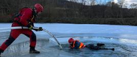 Zdjęcie przedstawia strażaków podczas ćwiczeń z zakresu ratownictwa lodowego na zamarzniętym stawie z wykorzystaniem węża pożarniczego napełnionego powietrzem