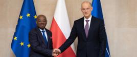 Na zdjęciu jest dwóch mężczyzn w ciemnych garniturach. Podają sobie dłonie i patrzą w obiektyw. Za nimi są trzy flagi: UE, Polski i NATO.