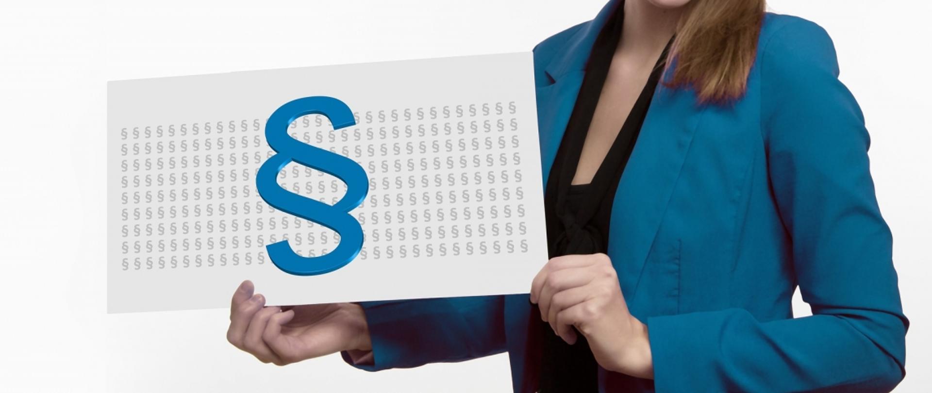 Kobieta w niebieskiej marynarce trzymająca planszę z dużym symbolem paragrafu (§). 