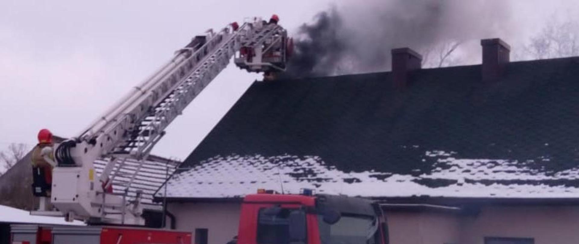 Pożar sadzy w kominie. Budynek jednopiętrowy, trzy kominy na dachu, z jednego wydobywa się czarny gęsty dym. Przed budynkiem rozstawiony samochód pożarniczy drabina. W koszu drabiny strażacy gaszący pożar sadzy w kominie