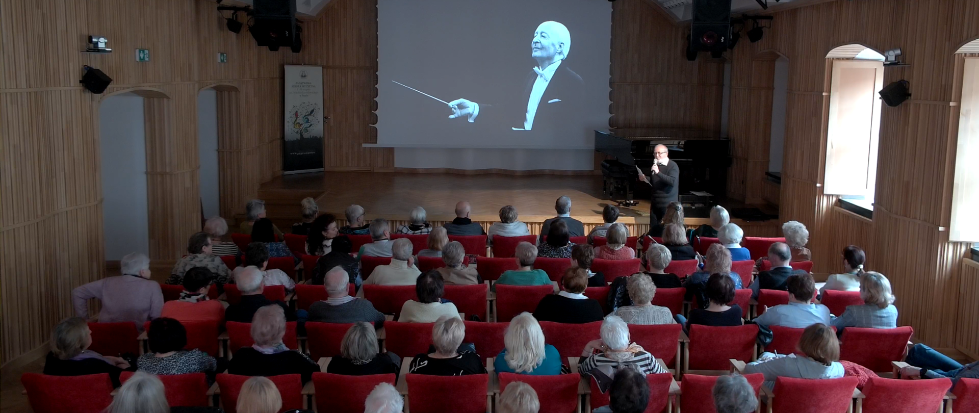 Zdjęcie. Sala koncertowa - widok od strony widowni. Na pierwszym planie widoczne od tylu rzędy słuchaczy. Przed nimi, tuż przed estradą stoi - zwrócony do publiczności - wykładowca trzymający mikrofon i kartkę. Za nim estrada, na której stoją z prawej strony dwa czarne fortepiany. Na ekranie wyświetlone jest zdjęcie dyrygującego kompozytora Witolda Lutosławskiego. W lewym rogu stoi baner PSM Nysa.