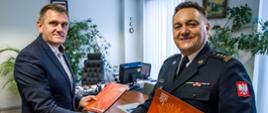 Dyrektor WORD Opole Pan Edward Kinder we własnym gabinecie ściska dłoń Opolskiego Komendanta Wojewódzkiego PSP w Opolu st. bryg. Krzysztofa Kędryka. Obaj Panowie trzymają w rękach czerwone ozdobne teczki PSP z wcześniej podpisanym porozumieniem.