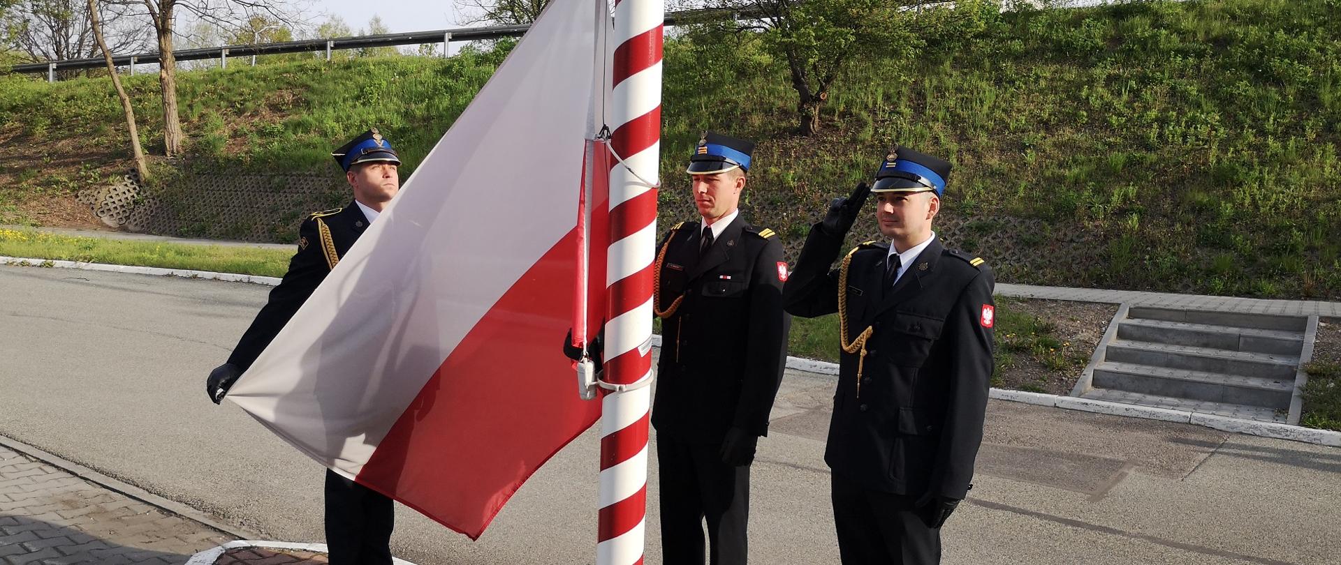 Zdjęcie przedstawia poczet flagowy w trakcie podnoszenia flagi państwowej. Poczet składa się z 3 strażaków.