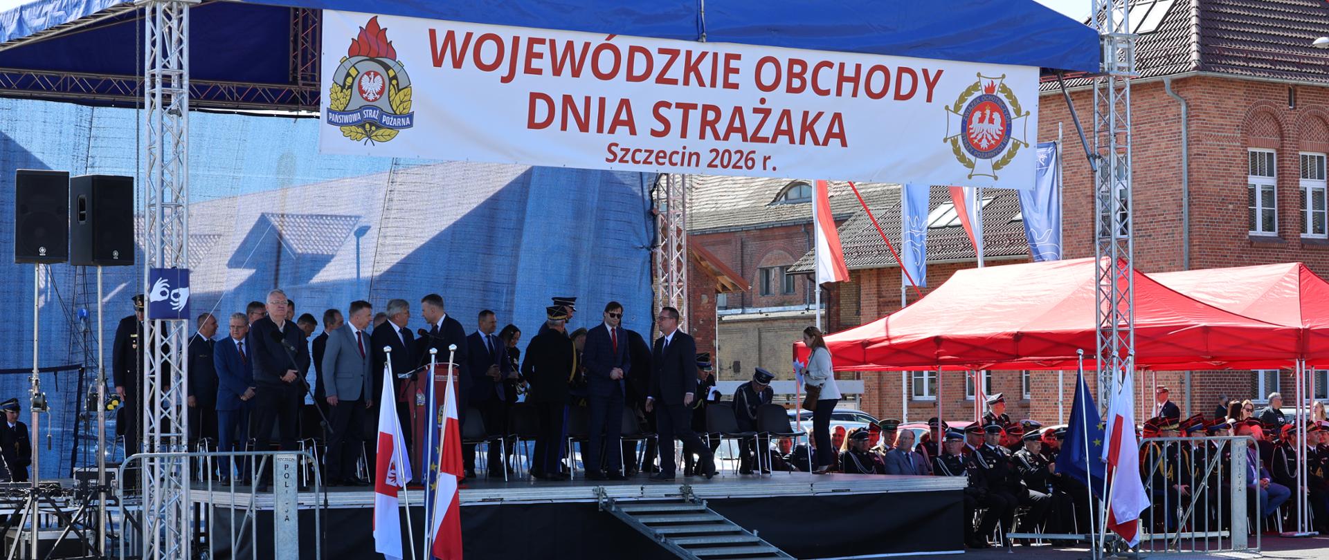 Uroczysta scena obchodów Dnia Strażaka w Szczecinie – pod zadaszeniem stoją strażacy i zaproszeni goście, obok widoczne są polskie i unijne flagi.