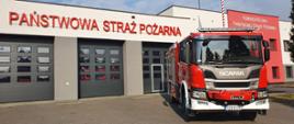 Samochód pożarniczy scania ustawiony przodem na tle garażu strażnicy. Po prawej stronie zielone drzewo iglaste. W tle na elewacji napis: Komenda Miejska Państwowej Straży Pożarnej.