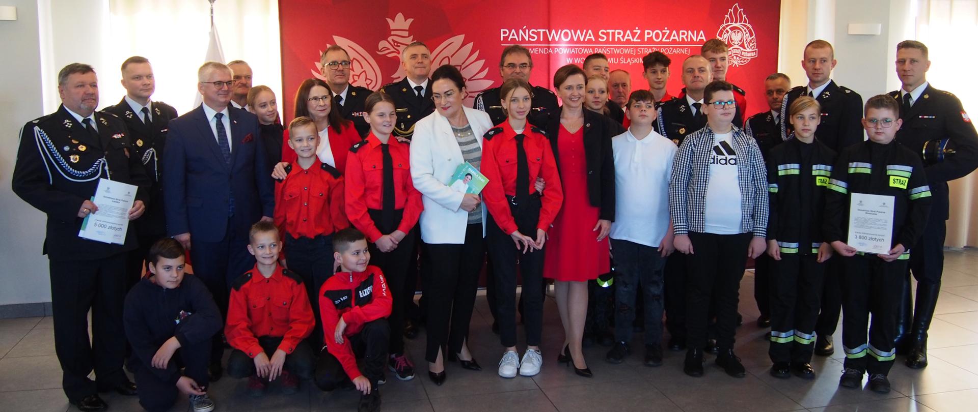 Strażacy w mundurach wyjściowych oraz dzieci i młodzież w mundurkach stoją na tle okien oraz ścianki z napisem Państwowa Straż Pożarna. 