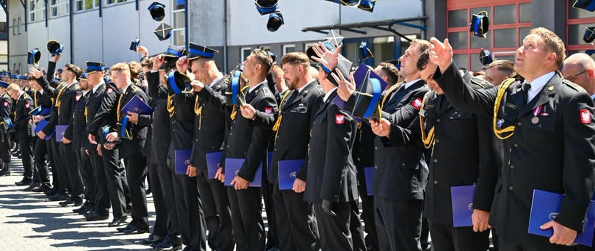 Grupa strażaków Państwowej Straży Pożarnej w galowych mundurach stoi w szeregu na placu przed budynkiem komendy. Wszyscy z uśmiechem i radością podrzucają w górę swoje czapki rogatywki podczas uroczystego apelu. W rękach trzymają niebieskie foldery lub dyplomy. W prawym dolnym rogu widnieje podpis: „st. str. kdt Piotr Krzak | Państwowa Straż Pożarna”.