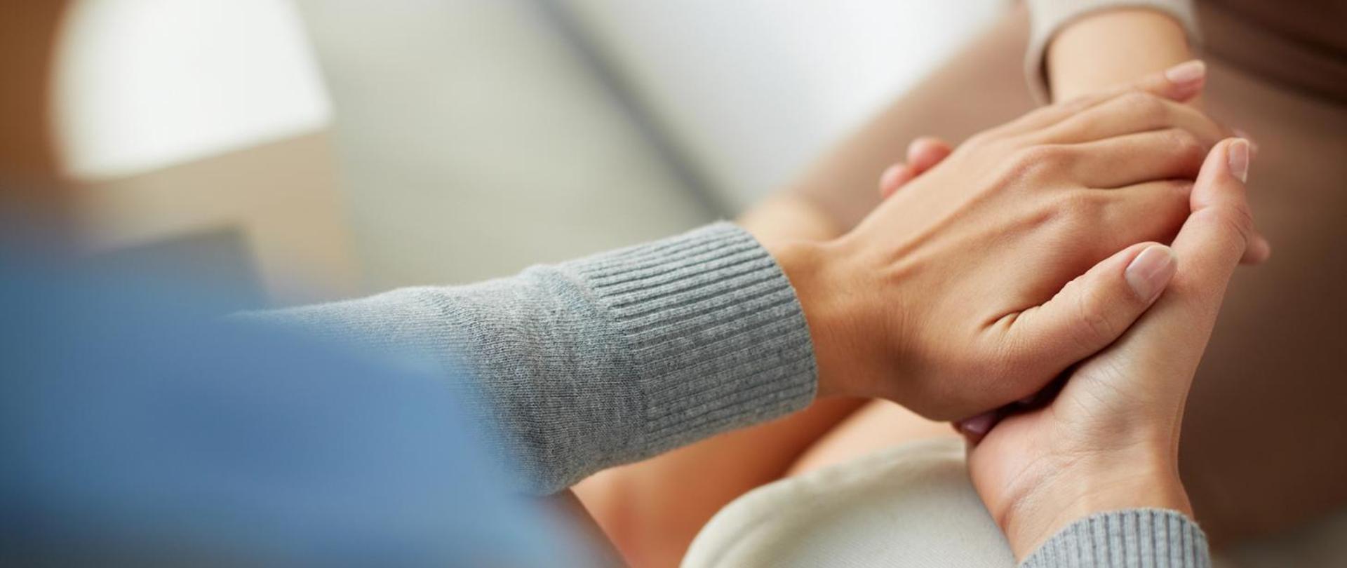 Dwie osoby trzymające się za ręce. Jedna osoba obejmuje dłoń drugiej w geście wsparcia, pocieszenia lub troski. Zdjęcie jest zbliżeniem na dłonie, tło jest rozmyte i jasne. Atmosfera wydaje się ciepła i pełna empatii.