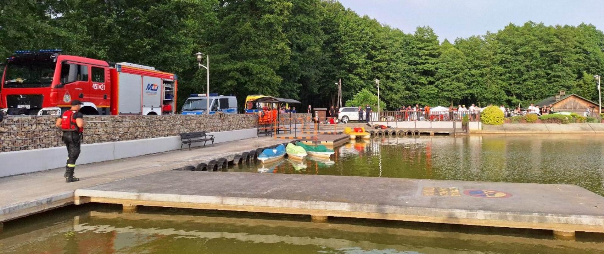 Na zdjęciu widoczny jest zalew ośrodka wypoczynkowego "Gutwin". Na pomoście widoczny jest funkcjonariusz Państwowej Straży Pożarnej. Na drugim planie widoczne są pojazdy Państwowej Straży Pożarnej, Policji oraz Pogotowia Ratunkowego.
