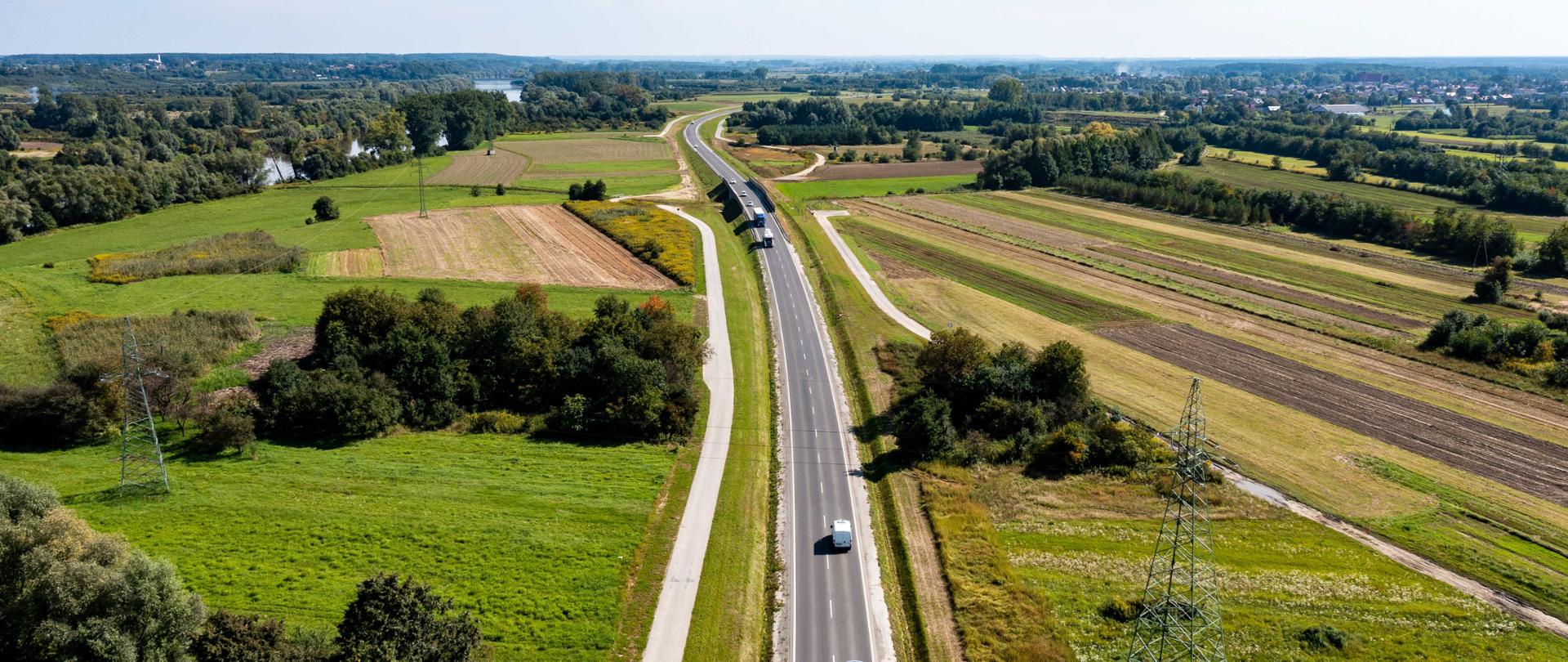 Widok z lotu ptaka na drogę przebiegającą przez rozległe tereny rolnicze. Po jezdni porusza się kilka samochodów, a po obu stronach drogi rozciągają się pola, łąki i skupiska drzew. W oddali widać rzekę oraz zabudowę. Nad krajobrazem rozciąga się jasne, lekko zachmurzone niebo.