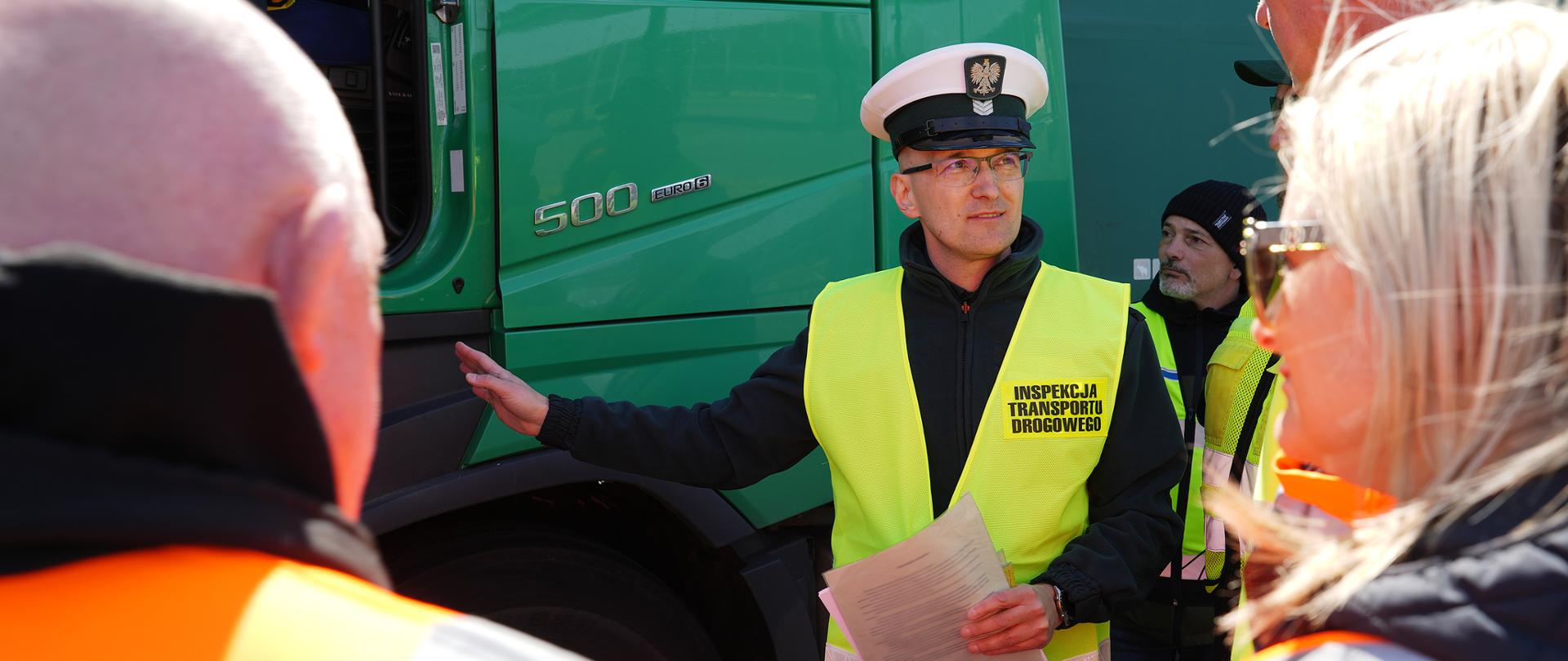 Funkcjonariusz w czapce i kamizelce odblaskowej stoi przy ciężarówce i wyjaśnia dokumenty grupie uczestników w trakcie warsztatów kontrolnych.