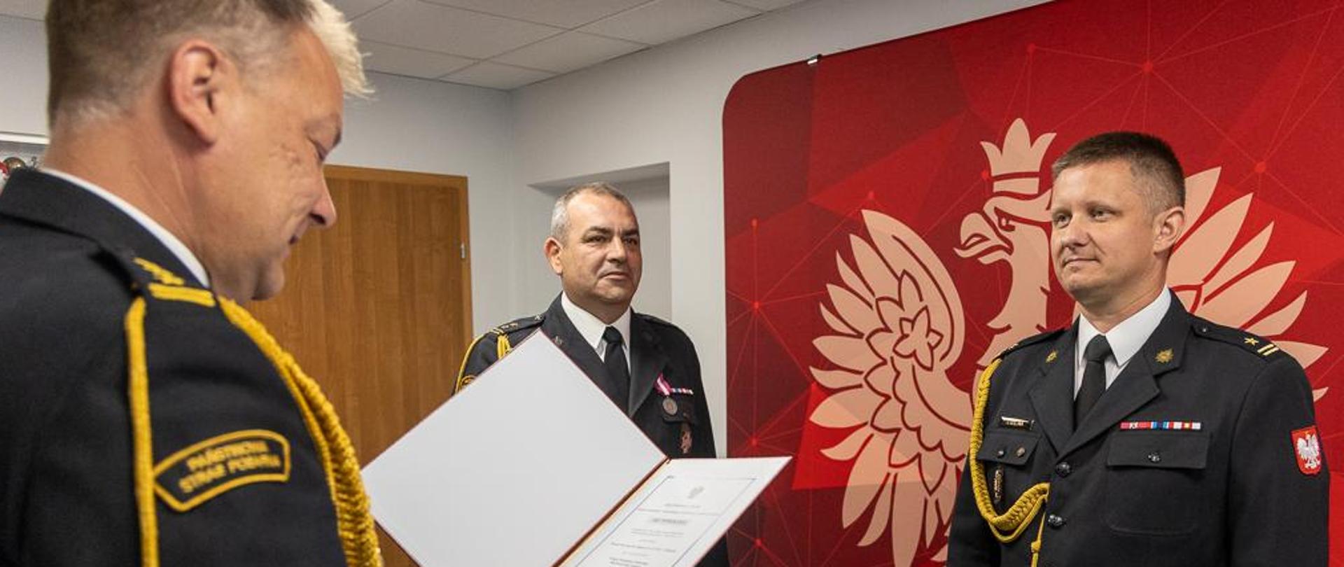 Na zdjęciu widać Opolskiego Komendanta Wojewódzkiego PSP odczytującego akt powołania Zastępcy Komendanta Powiatowego PSP w Kluczborku a także Komendanta Powiatowego PSP w Kluczborku - wszyscy w umundurowaniu galowym na tle ścianki z logotypem Państwowej Straży Pożarnej.