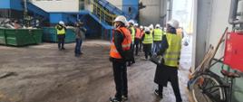 Osoby w kamizelkach odblaskowych i kaskach stoją w dużym, magazynowym pomieszczeniu.