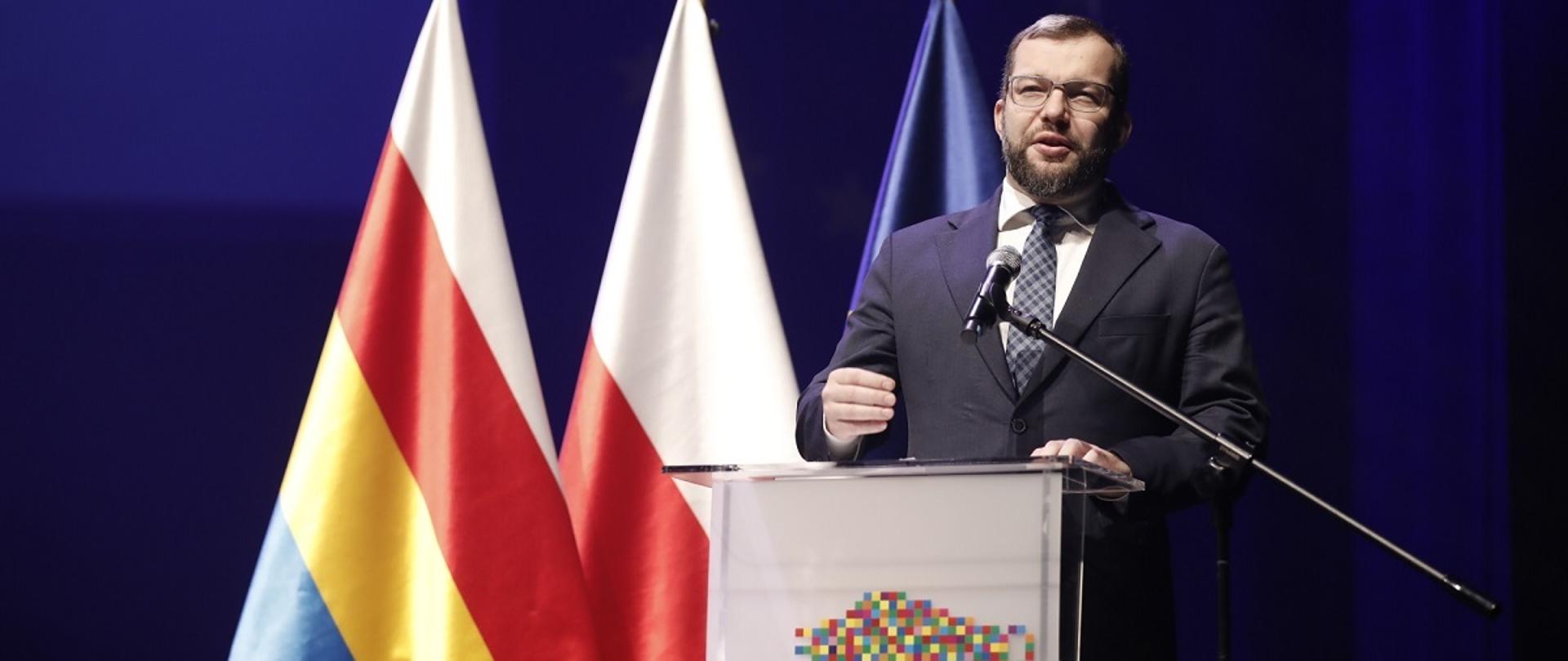 Minister funduszy i polityki regionalnej Grzegorz Puda podczas inauguracji Funduszy Europejskich dla Podlasia w Białymstoku, minister stoi przy mównicy, za nim flagi Polski, UE i Podlasia