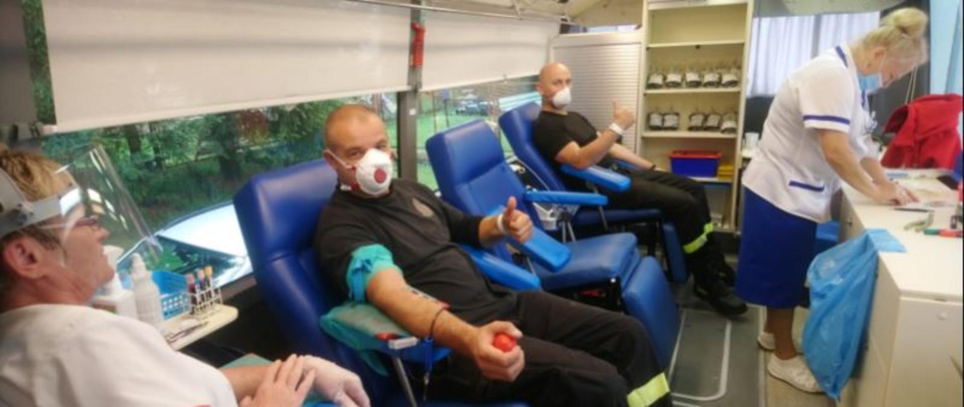 Zdjęcie przedstawia dwóch strażaków oddających krew w specjalnym autobusie. Funkcjonariusze mają na twarzy maseczki. W autobusie znajdują się także dwie pielęgniarki, które prowadzą i nadzorują akcję poboru krwi.