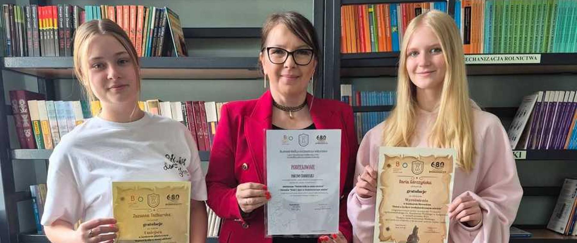 Zdjęcie przedstawia trzy stojące obok siebie kobiety w bibliotece szkolnej, trzymające pamiątkowe dyplomy.Postacie od lewej do prawejMłoda kobieta po lewej: Ma jasne, spięte z tyłu włosy i luźne ubranie. Nosi biały t-shirt oraz szare, dresowe spodnie. W prawej ręce trzyma ozdobny, żółtawy dyplom.Kobieta w środku: Jest starsza od pozostałych osób, nosi okulary w ciemnych oprawkach i ma ciemne włosy spięte z tyłu. Ubrana jest w elegancką, czerwoną marynarkę, czarną bluzkę i czarne, szerokie spodnie. Prezentuje biały dyplom w foliowej koszulce.Młoda kobieta po prawej: Ma długie, proste, blond włosy. Jest ubrana w jasnoróżową, luźną bluzę oraz ciemne spodnie. W dłoniach trzyma ozdobny dyplom o stylizowanym, żółtawym odcieniu.Otoczenie i tłoRegały z książkami: Za kobietami stoją wysokie, ciemne regały biblioteczne wypełnione książkami o kolorowych okładkach. Widoczne są białe napisy działów, m.in. „LEKTURY” oraz „MECHANIZACJA ROLNICTWA”.Logo szkoły: Po prawej stronie, na dolnej części regału, umieszczono kwadratowe logo z rysunkiem kłosa zboża, ryby oraz napisem „ZS KOWAL”.Podłoże i oświetlenie: Kobiety stoją na jasnych, ukośnie ułożonych płytkach podłogowych. Światło pada z przodu, wyraźnie oświetlając postacie.