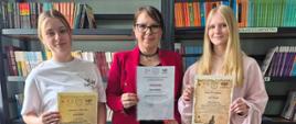 Zdjęcie przedstawia trzy stojące obok siebie kobiety w bibliotece szkolnej, trzymające pamiątkowe dyplomy.Postacie od lewej do prawejMłoda kobieta po lewej: Ma jasne, spięte z tyłu włosy i luźne ubranie. Nosi biały t-shirt oraz szare, dresowe spodnie. W prawej ręce trzyma ozdobny, żółtawy dyplom.Kobieta w środku: Jest starsza od pozostałych osób, nosi okulary w ciemnych oprawkach i ma ciemne włosy spięte z tyłu. Ubrana jest w elegancką, czerwoną marynarkę, czarną bluzkę i czarne, szerokie spodnie. Prezentuje biały dyplom w foliowej koszulce.Młoda kobieta po prawej: Ma długie, proste, blond włosy. Jest ubrana w jasnoróżową, luźną bluzę oraz ciemne spodnie. W dłoniach trzyma ozdobny dyplom o stylizowanym, żółtawym odcieniu.Otoczenie i tłoRegały z książkami: Za kobietami stoją wysokie, ciemne regały biblioteczne wypełnione książkami o kolorowych okładkach. Widoczne są białe napisy działów, m.in. „LEKTURY” oraz „MECHANIZACJA ROLNICTWA”.Logo szkoły: Po prawej stronie, na dolnej części regału, umieszczono kwadratowe logo z rysunkiem kłosa zboża, ryby oraz napisem „ZS KOWAL”.Podłoże i oświetlenie: Kobiety stoją na jasnych, ukośnie ułożonych płytkach podłogowych. Światło pada z przodu, wyraźnie oświetlając postacie.
