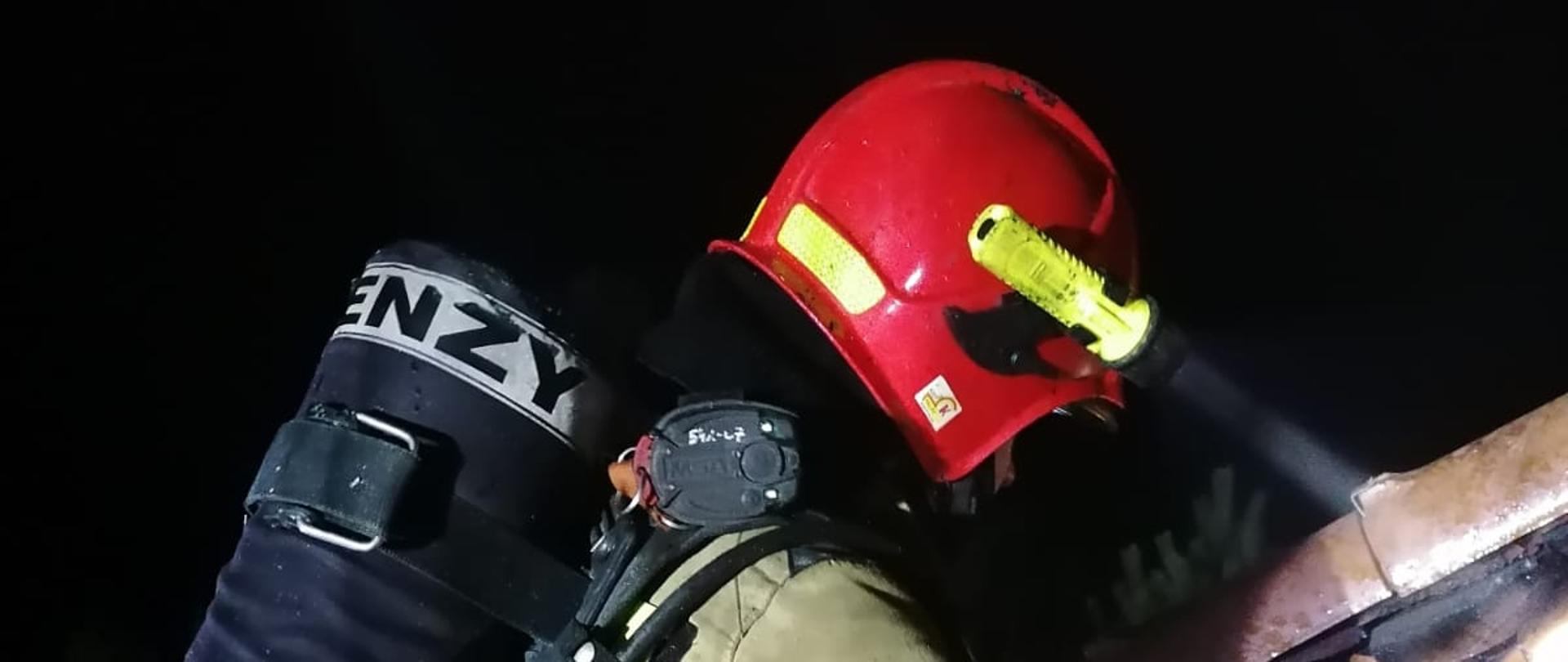 Strażak w pozycji kucznej jest na dachu. Ubrany w ubranie specjalne i hełm. Na plecach ma butlę z powietrzem. Z przyczepionej do hełmu latarki wydobywa się światło. Wokół strażaka jest ciemno. 