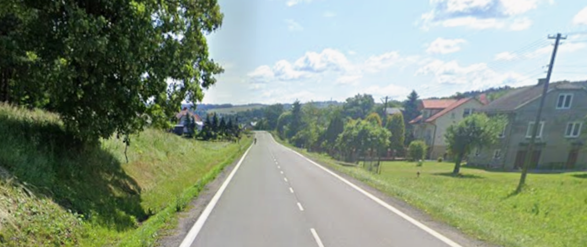 Na środku zdjęcia widać drogę, po obu stronach brak jest chodnika, jest pobocze, po prawej stronie widać domu.
