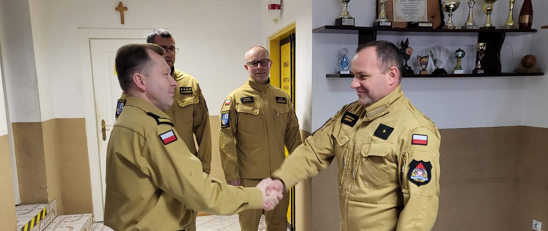 Starszy brygadier Maciej Suchomski składa gratulacje awansowanemu strażakowi, młodszy brygadier Dariusz Chylewski oraz starszy kapitan Krzysztof Brzozowski ubrani w mundury służbowe zgromadzeni w pomieszczeniu świetlicy Komendy Powiatowej Państwowej Straży Pożarnej w Sępólnie Krajeńskim. 
