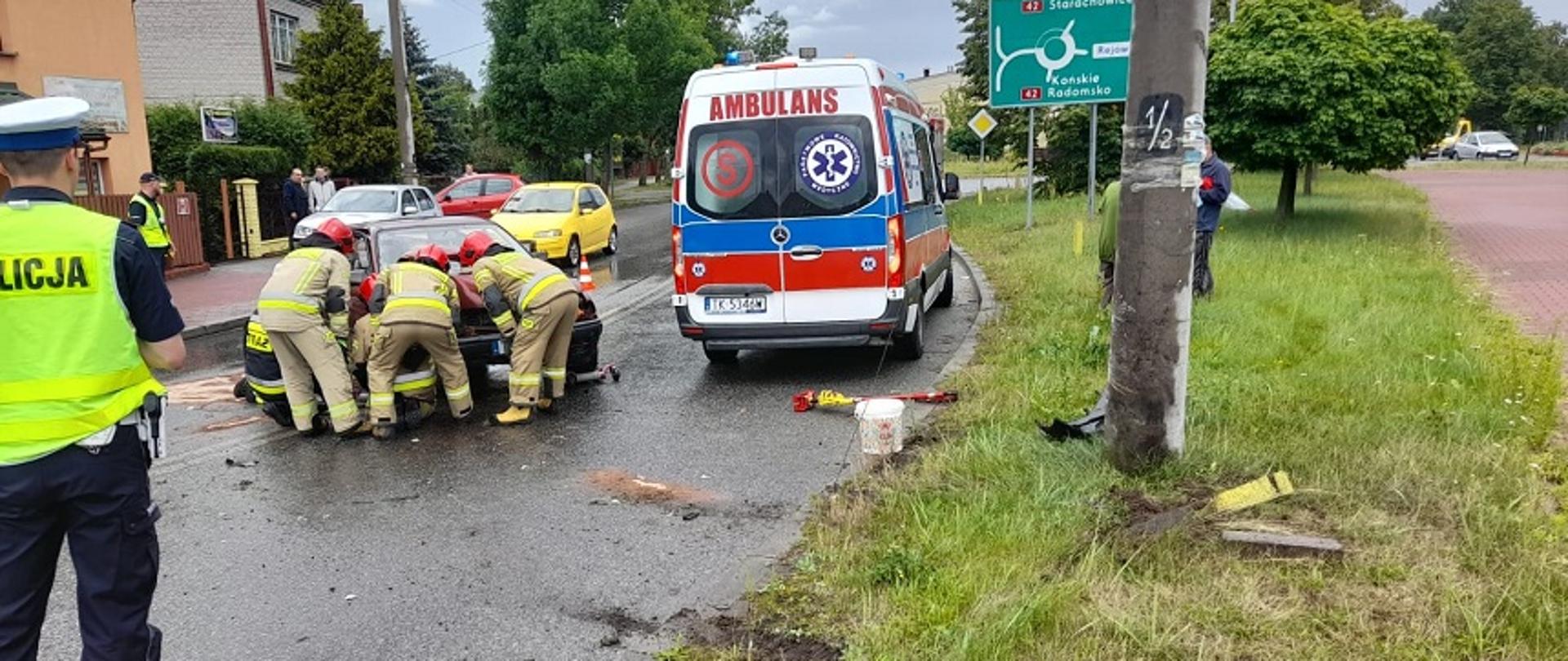 Zdjęcie obrazuje samochód osobowy, który uderzył w przydrożny słup energetyczny. W wyniku uderzenia został on odrzucony na środek drogi. Wokół niego znajdują się strażacy, którzy podnoszą maskę samochodu celem odłączenia akumulatora w rozbitym pojeździe. Z prawej strony widać samochód Zespołu Ratownictwa Medycznego.