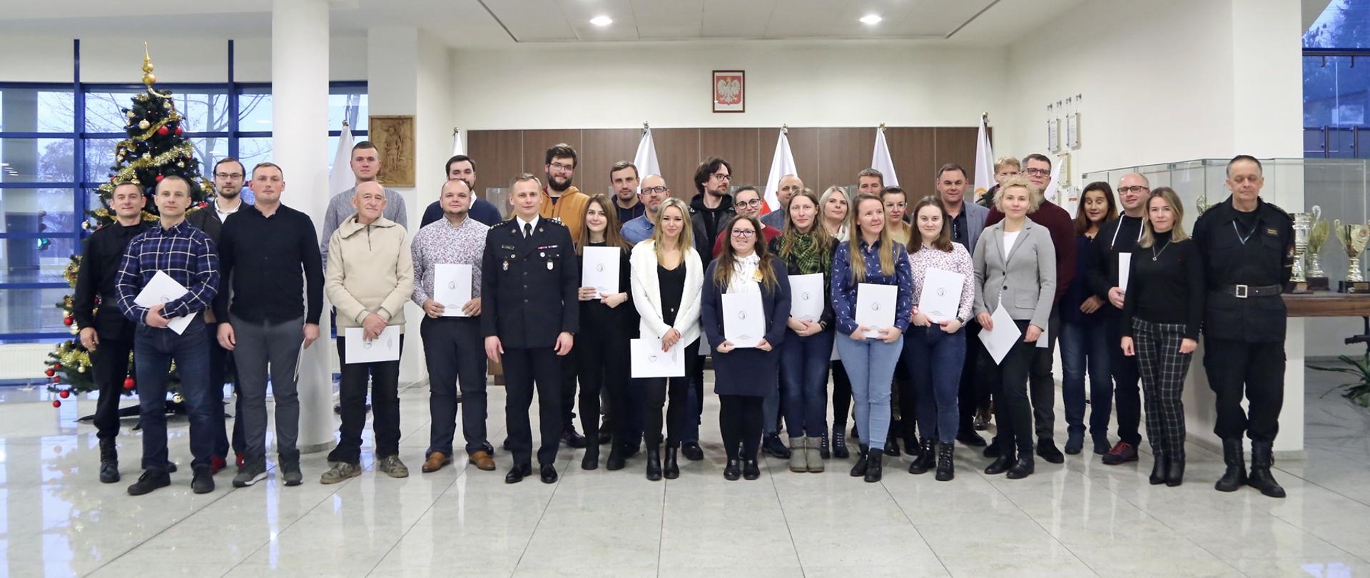 Grupowe zdjęcie uczestników szkolenia (28 osób) w towarzystwie Zastępcy Komendanta Centralnej Szkoły PSP, dwóch funkcjonariuszy CS PSP oraz kobiety. Uczestnicy szkolenia trzymają otrzymane zaświadczenia