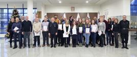 Grupowe zdjęcie uczestników szkolenia (28 osób) w towarzystwie Zastępcy Komendanta Centralnej Szkoły PSP, dwóch funkcjonariuszy CS PSP oraz kobiety. Uczestnicy szkolenia trzymają otrzymane zaświadczenia