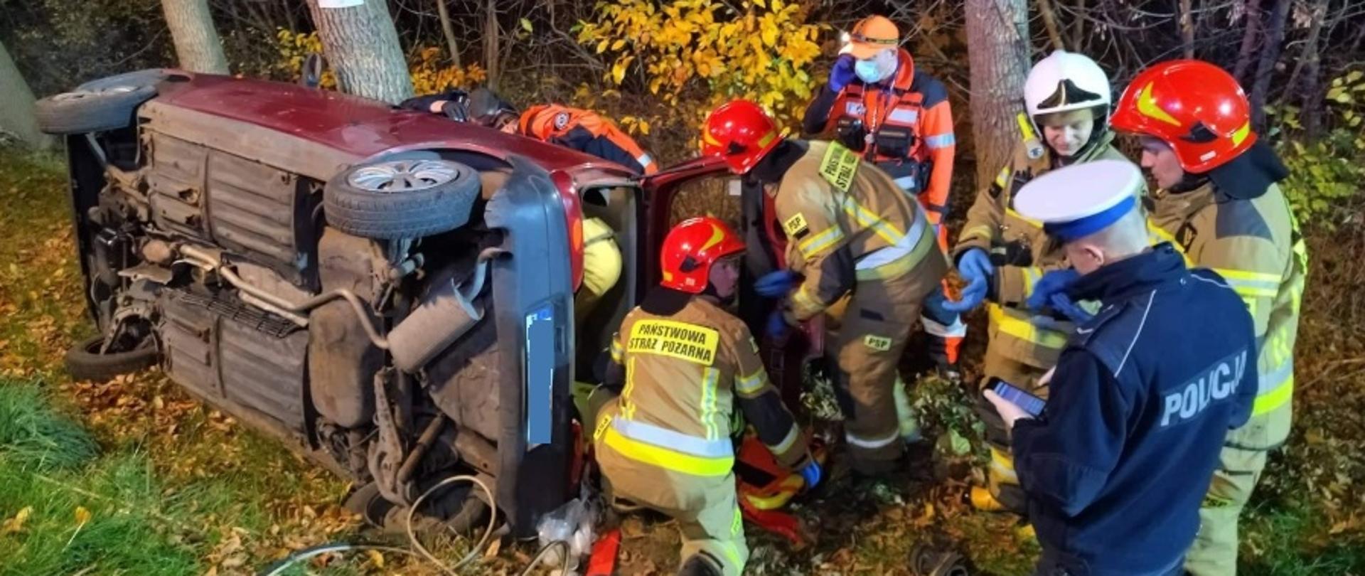 Na zdjęciu widać samochód osobowy koloru bordowego, leżący na prawym boku. Wokół pojazdu widoczni są strażacy, ratownicy medyczni oraz policjant.