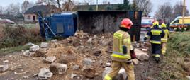 Strażacy przechodzą przy samochodzie ciężarowym, który leży na boku. Na jezdni rozsypany gruz.