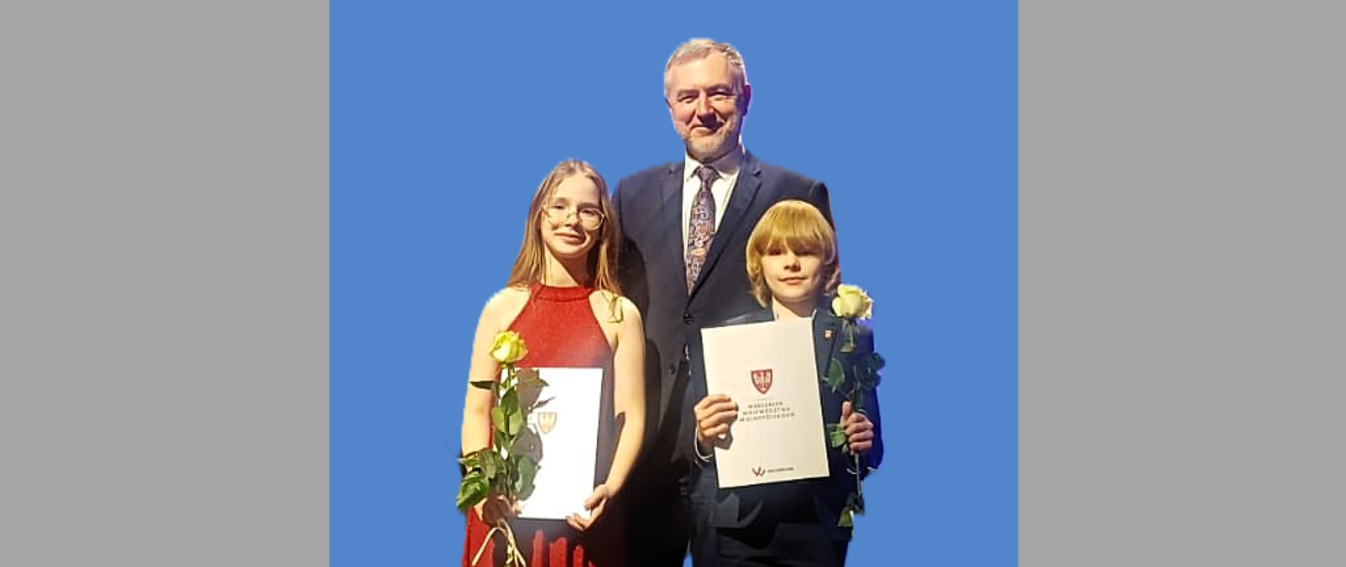 dwoje dzieci trzymających dyplomy i kwiaty, za nimi mężczyzna w garniturze, niebieskie tło