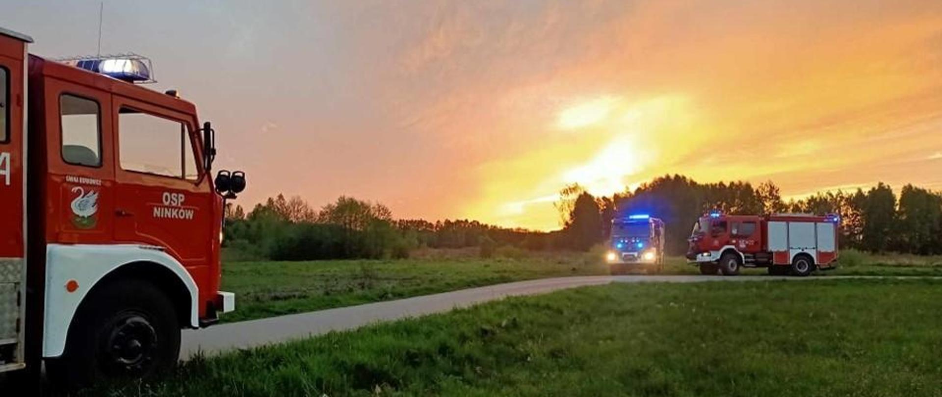 Trzy wozy strażackie stoją na polnej drodze o zachodzie słońca, z czerwonym niebem w tle. Na pierwszym planie widać fragment czerwonego wozu strażackiego z napisem "OSP NINKÓW" i włączonymi niebieskimi światłami. Drugi wóz ma włączone światła drogowe, a trzeci jest częściowo widoczny w oddali.