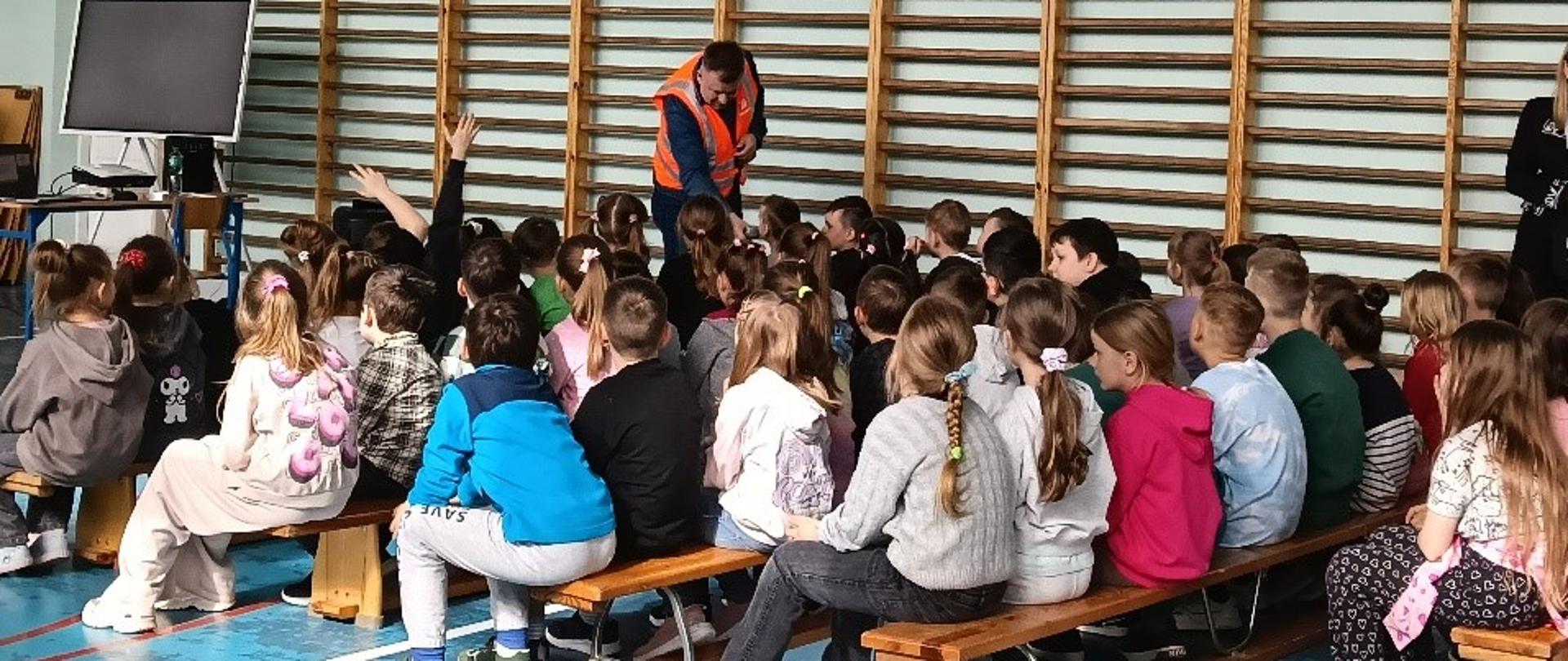 Zdjęcie przedstawia zajęcia edukacyjne odbywające się w sali gimnastycznej. Widoczna jest grupa dzieci siedzących na ławkach oraz osoba prowadząca spotkanie. W tle znajdują się elementy wyposażenia sali sportowej.