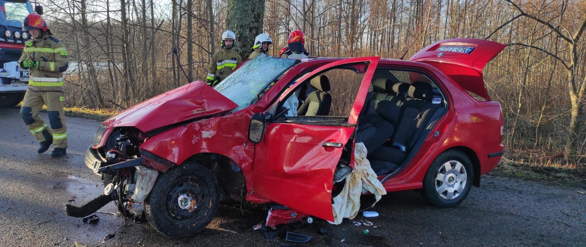 Czerwone auto osobowe, rozbite, uszkodzone po uderzeniu w drzewo, otwarte drzwi i bagażnik. Przy nim strażacy.