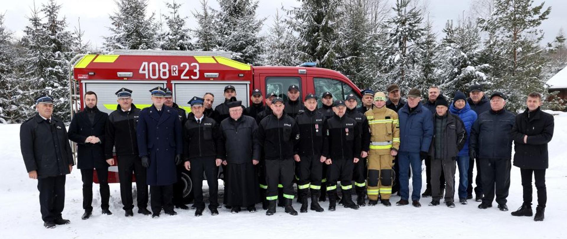 aNa zdjęciu widać wszystkich obecnych na przekazaniu samochodu ratowniczego w OSP Wojtkówce. W tym komendant powiatowy PSP w Ustrzykach Dolnych mł. bryg. Emil Szmyd, druh Tadeusz Niedżwiecki, burmistrz miasta i gminy Ustrzyki Dolne Pan Bartosz Romowicz, Zastępca burmistrza Pan Michał Wnuk, ksiądz, druhowie OSP Wojtkówka oraz przedstawiciele władz. Za wszystkimi widać nowy samochód VW Crafter. Aura na zdjęciu jest zimowa i w około widać drzewa.