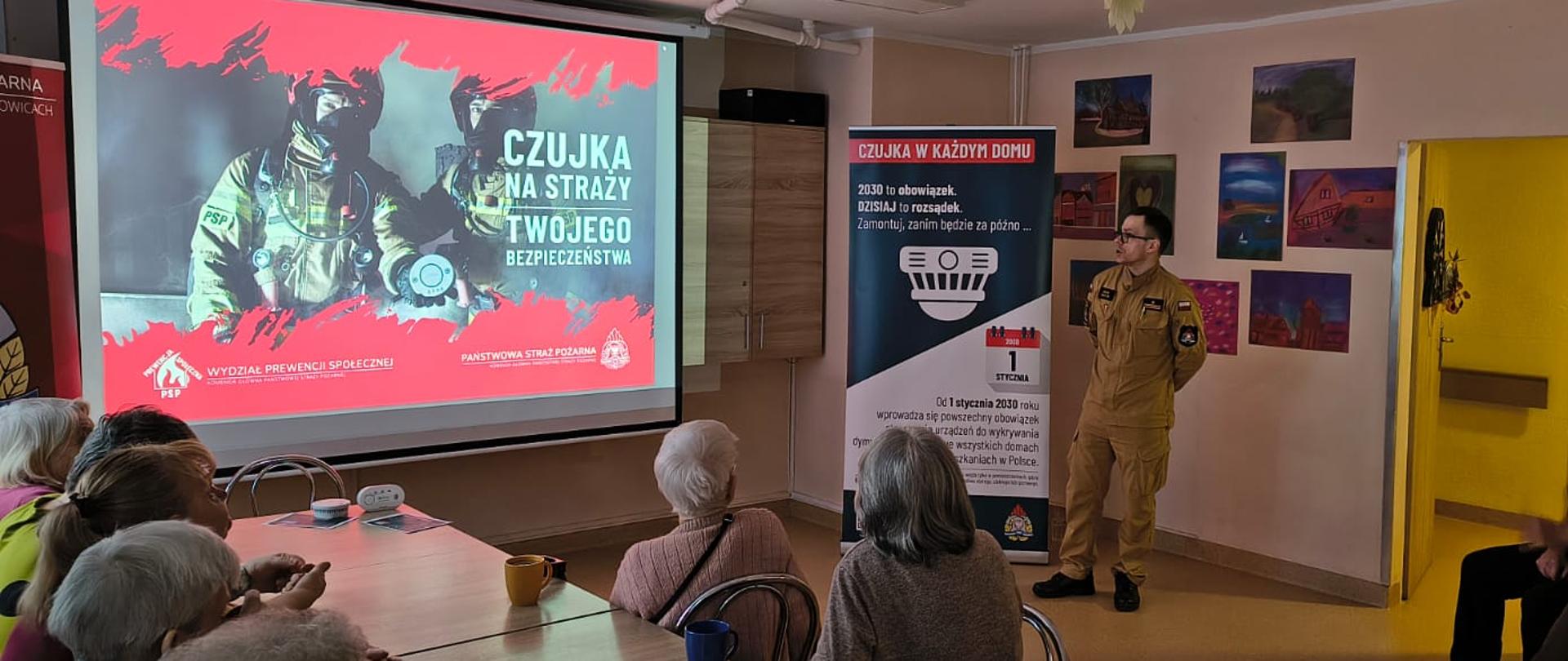 Na zdjęciu uwieczniono spotkanie edukacyjne prowadzone przez funkcjonariuszy Państwowej Straży Pożarnej w Świętochłowicach. Jeden strażak w piaskowym mundurze służbowym prezentuje materiały szkoleniowe grupie seniorów siedzących przy okrągłych stolikach. W tle widoczny jest ekran z prezentacją dotyczącą sygnałów alarmowych oraz baner informacyjny kampanii „Czujka na straży Twojego Bezpieczeństwa”, co sugeruje, że tematyka spotkania dotyczy szeroko pojętego bezpieczeństwa przeciwpożarowego i obrony cywilnej.