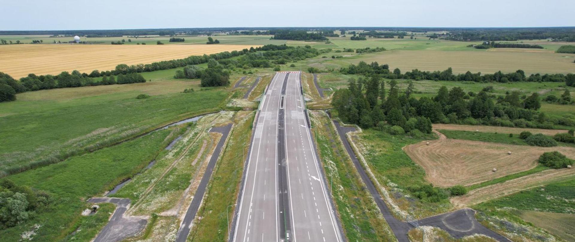 Widok z lotu ptaka na nowo wybudowany odcinek autostrady A2 przebiegający przez tereny rolnicze w okolicach Białej Podlaskiej. Dwujezdniowa droga biegnie przez zielone pola i zadrzewienia, a po obu stronach widoczne są rowy odwadniające oraz drogi techniczne.
