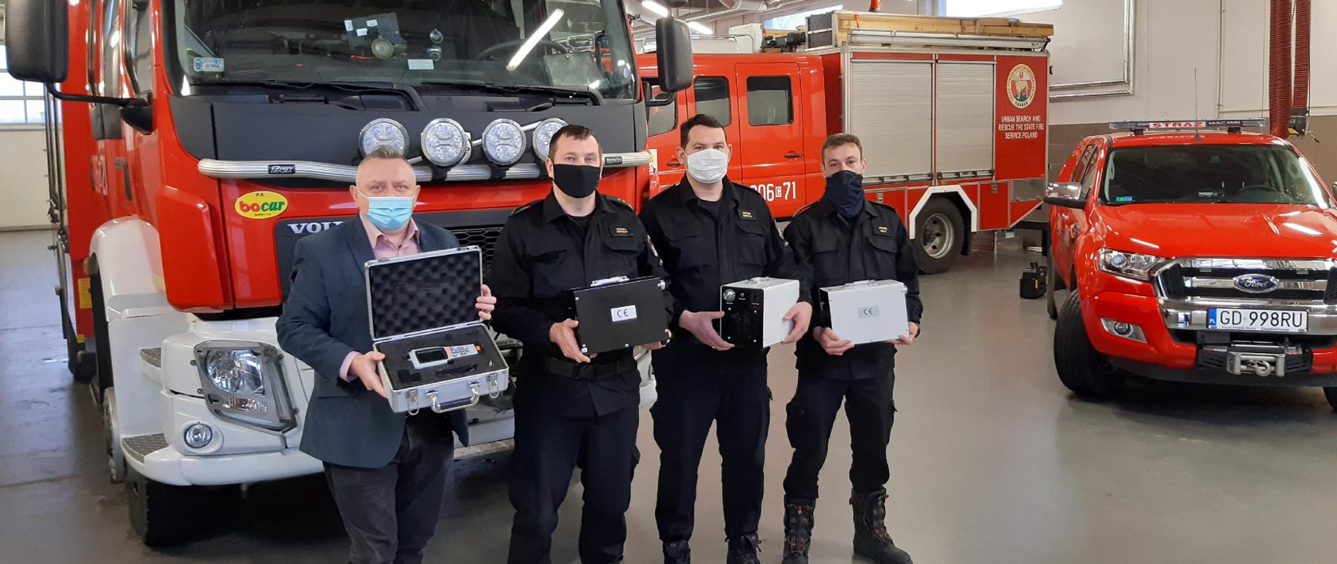 Na pierwszym planie Piotr Lachowski - Przewodniczący Rady Dzielnicy Ujeścisko-Łostowice, następnie bryg. Maciej Sapieha - Dowódca JRG 2 i dwóch strażaków. Wszyscy trzymają sprzęt przekazany od Rady Dzielnicy. Sprzęt to generatory ozonu wraz z specjalistycznym miernikiem. W tle samochody pożarnicze w garażu Posterunku JRG 2.