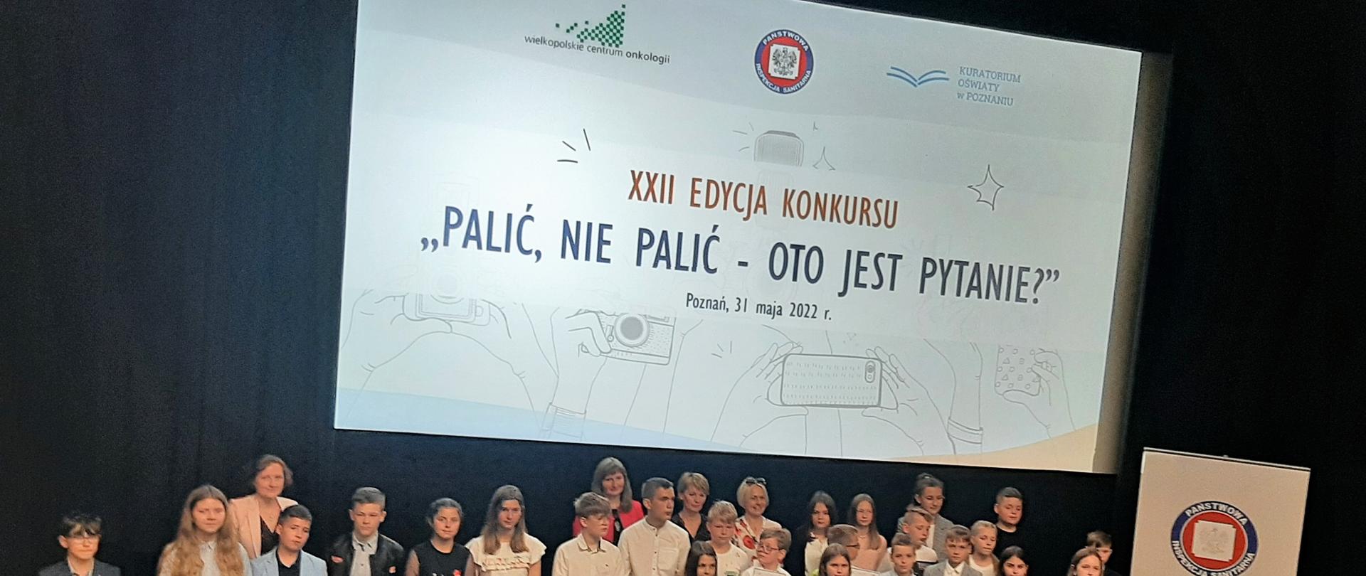 1. Na zdjęciu widać grupę dzieci – laureatów konkursu „Palić, nie palić – oto jest pytanie?” w sali kinowej ustawionych przodem pod ekranem, w tle za dziećmi widoczna jest plansza z napisem „XXII edycja konkursu „Palić, nie palić – oto jest pytanie?”