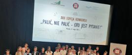 1. Na zdjęciu widać grupę dzieci – laureatów konkursu „Palić, nie palić – oto jest pytanie?” w sali kinowej ustawionych przodem pod ekranem, w tle za dziećmi widoczna jest plansza z napisem „XXII edycja konkursu „Palić, nie palić – oto jest pytanie?”
