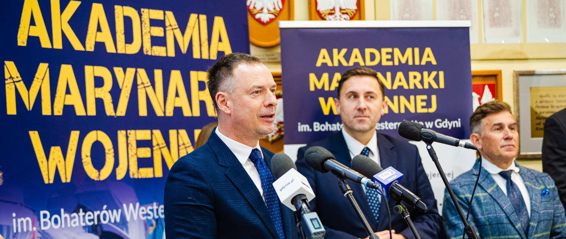 Piotr Borys podczas konferencji w Gdyni. Stoi za mikrofonem na tle ścianki z logiem Akademii Marynarki Wojennej