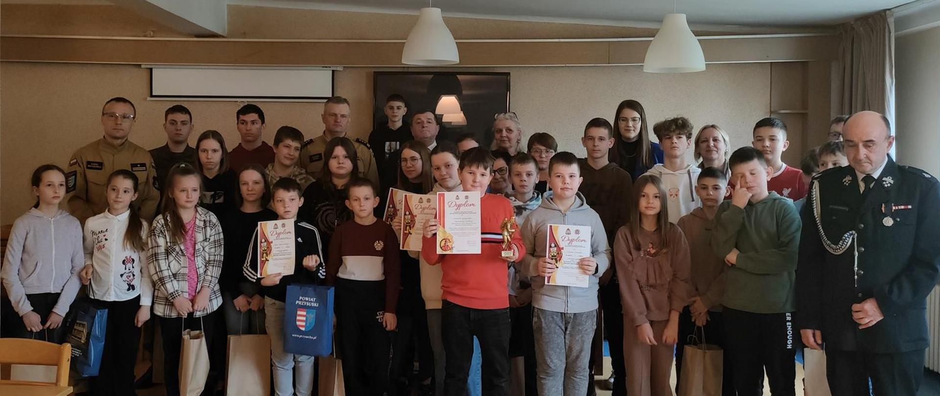 Zdjęcie przedstawia grupę osób, w tym dorosłych i dzieci, stojących razem w pomieszczeniu, prawdopodobnie sali konferencyjnej lub wykładowej. Po prawej stronie widać starszego mężczyznę w mundurze strażackim, a w głębi po lewej stronie kilku innych dorosłych mężczyzn, również w mundurach, oraz kobietę.
Większość osób na zdjęciu to dzieci i młodzież. Niektóre z nich trzymają w rękach dyplomy i trofea, co sugeruje uroczystość wręczenia nagród lub zakończenia konkursu. Na przykład, chłopiec w czerwonym swetrze w centrum kadru trzyma złotą statuetkę i dyplom. Inny chłopiec obok niego również trzyma dyplom. Dzieci są ubrane w codzienne ubrania.
W tle widoczne są ściany, a na suficie wiszą białe lampy. Po lewej stronie widać fragment stołu. Na dole, w centrum kadru, leży niebieska torba z napisem "POWIAT PODDĘBICKI". Całość obrazu przedstawia uroczystą chwilę, być może zakończenie zawodów lub kursu związanego ze strażą pożarną.