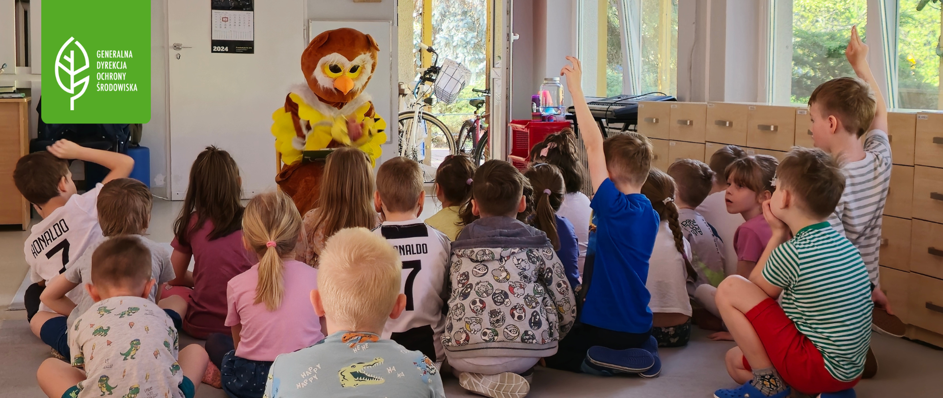 W rzędach siedzą dzieci w wieku przedszkolnym, postać przebrana za sowę siedzi na krześle. Część dzieci podnosi rączkę do góry.