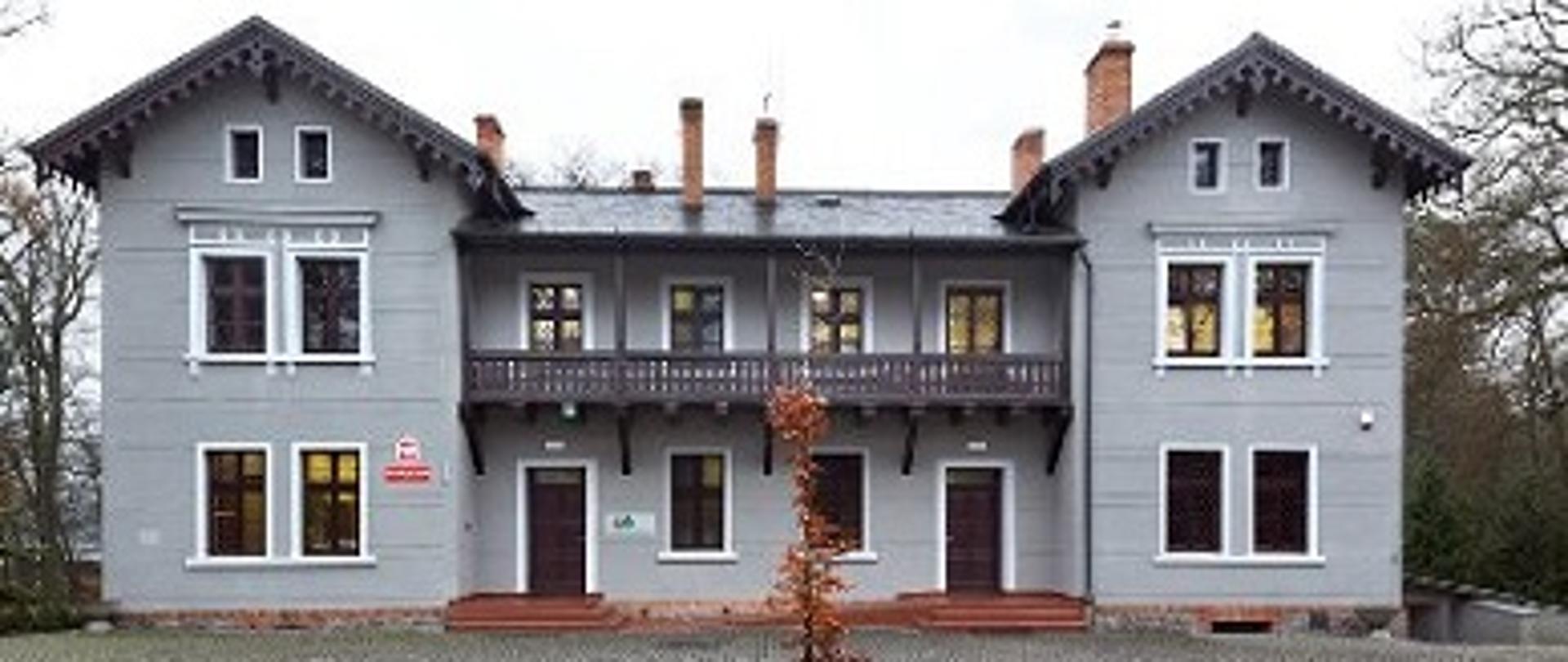 Na środku zdjęcia stoi szary piętrowy budynek z balkonem oraz dwoma wejściami do których prowadzą niskie schody, z przodu budynku rośnie drzewo, podwórko wyłożone kostką granitową.