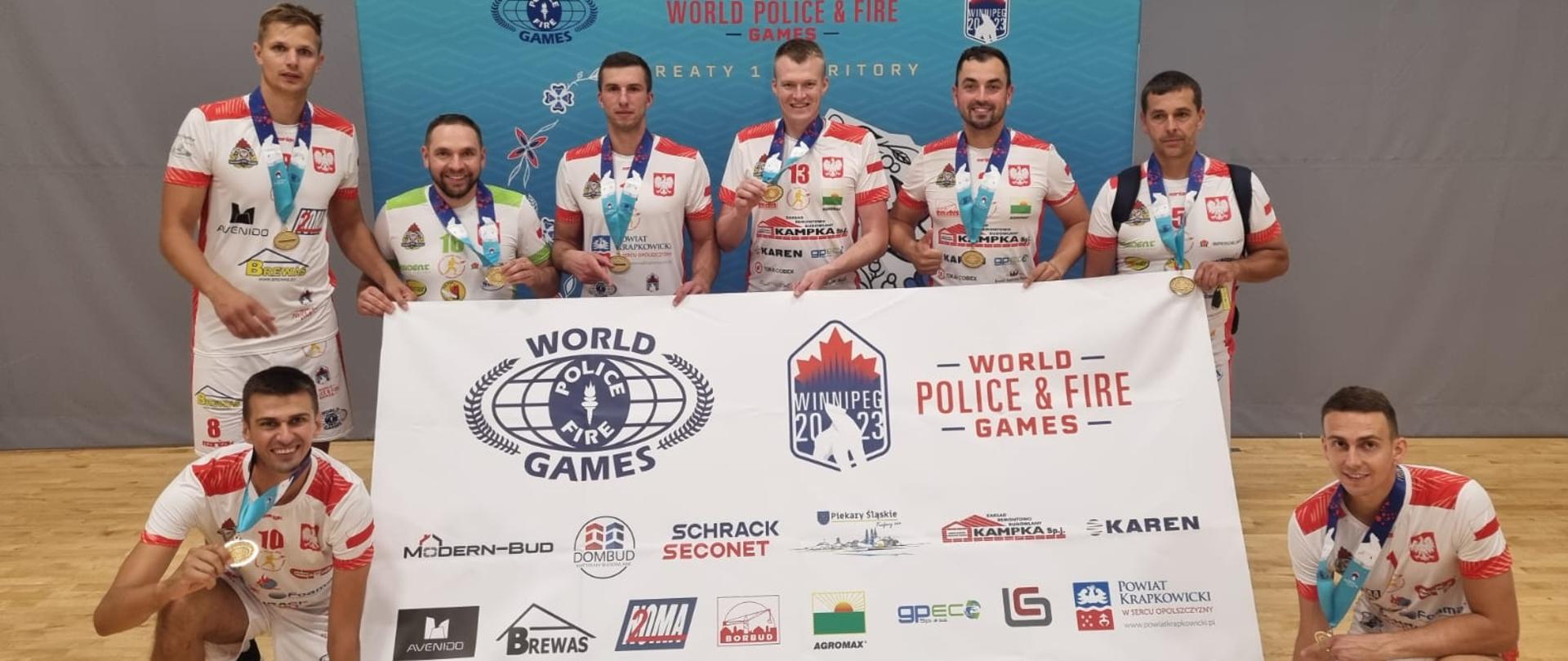Zawodnicy w strojach sportowych z medalem na szyi stojący wokół trzymanego baneru z nazwą imprezy, czyli World Police and Fire Games oraz umieszczonymi poniżej nazwami sponsorów