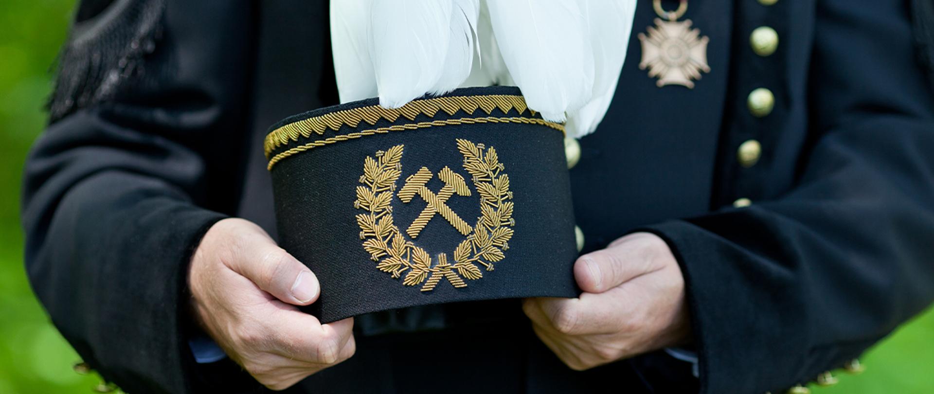 Bliskie ujęcie górnika w galowym stroju: widoczna czapka z białym pióropuszem, trzymające ją dłonie i część góry munduru.