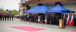 Na zdjęciu widoczna trybuna honorowa oraz zaproszeni goście strażacy PSP, OSP, parlamentarzyści, samorządowcy pod niebieskim namiotem. Po lewej stronie poczty sztandarowe OSP z powiatu gorlickiego. Po lewej stronie mównica i przemawiający senator RP.