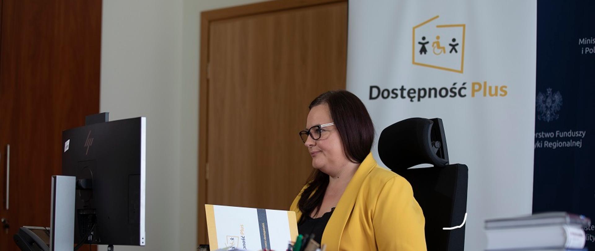 Minister Jarosińska-Jedynak siedzi przy biurku, patrzy w ekran komputera. Na biurku widoczny stos dokumentów, w tle ścianka z logo programu Dostępność Plus.